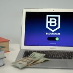 Laptop exibindo uma tela conceitual de conexão blockchain sobre uma mesa com livros e cédulas de dólar.