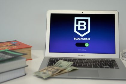 Laptop exibindo uma tela conceitual de conexão blockchain sobre uma mesa com livros e cédulas de dólar.