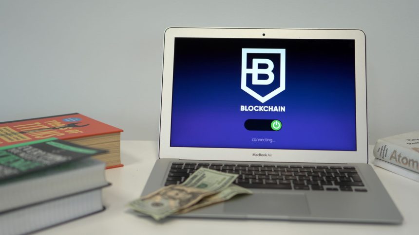 Laptop exibindo uma tela conceitual de conexão blockchain sobre uma mesa com livros e cédulas de dólar.