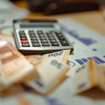Cédulas de euro e calculadora sobre documentos financeiros, em uma composição que remete a orçamento e contabilidade.