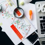 Documentos financeiros com gráficos e um laptop organizados sobre uma mesa de trabalho.