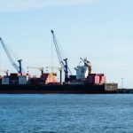 Navio cargueiro carregado de contêineres atracado em um porto comercial, ao lado de guindastes.