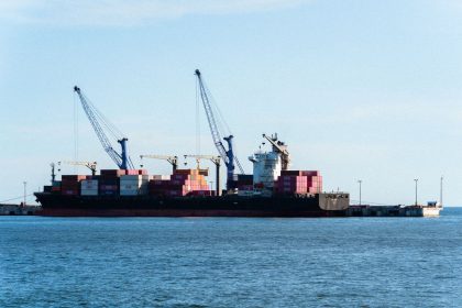 Navio cargueiro carregado de contêineres atracado em um porto comercial, ao lado de guindastes.