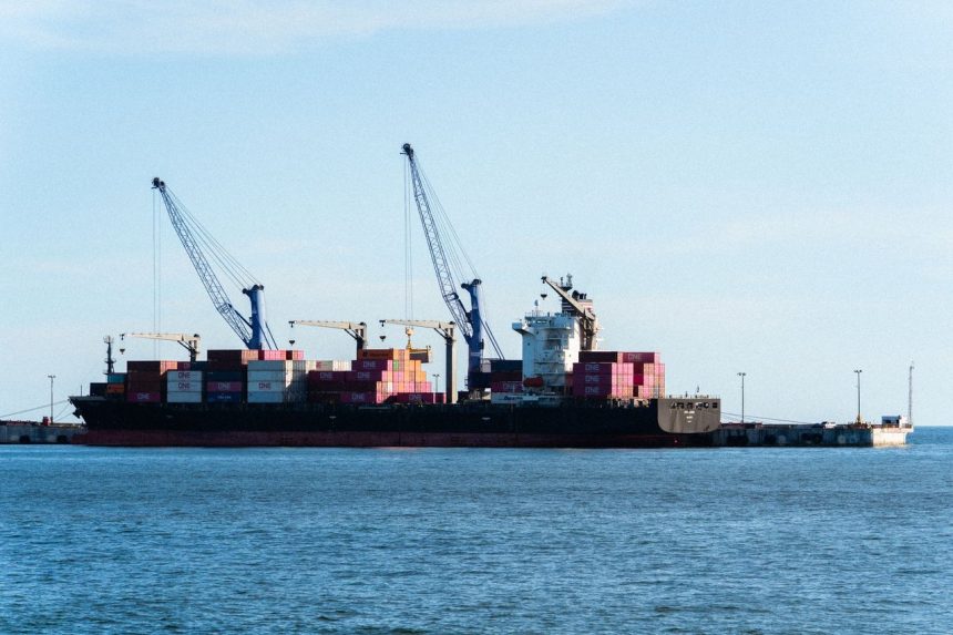 Navio cargueiro carregado de contêineres atracado em um porto comercial, ao lado de guindastes.
