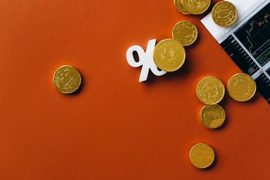 Moedas douradas sobre um gráfico do mercado financeiro, acompanhadas de um símbolo de porcentagem.